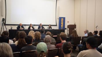 A panel discussion with four speakers at a university event, audience members in foreground.